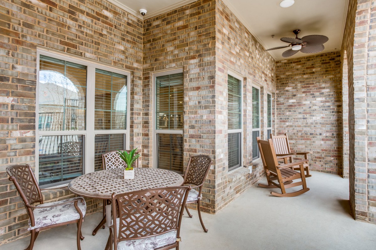 A cozy patio with brick walls featuring a table and chairs, along with rocking chairs under a ceiling fan.