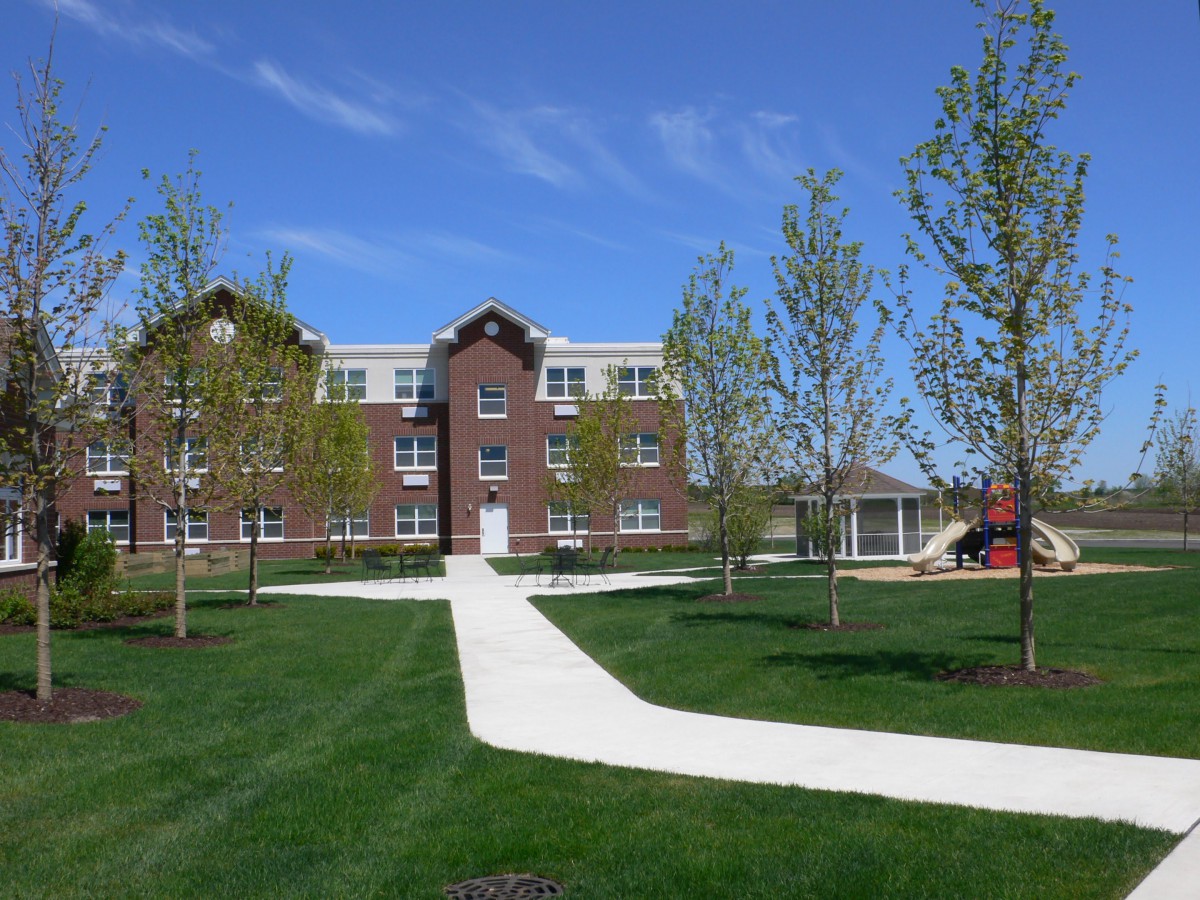 A brick building with a white pathway surrounded by greenery and trees under a clear blue sky.
