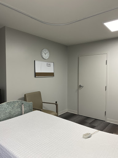 A modern hospital room featuring a bed, chair, and a wall clock.