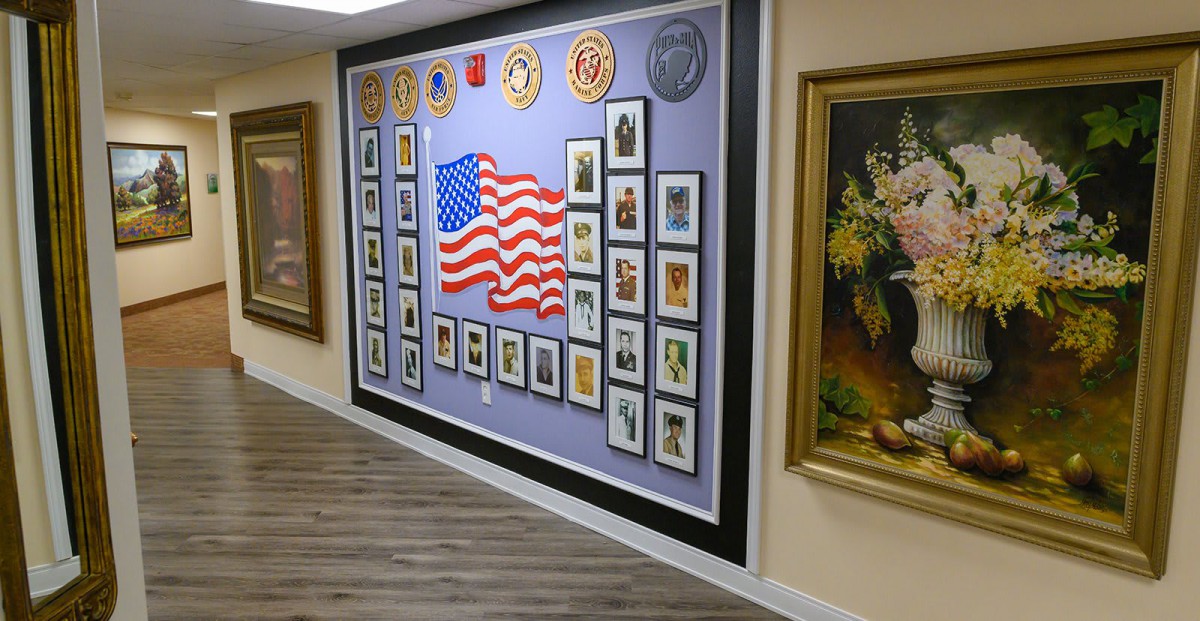 A decorated wall featuring an American flag surrounded by military emblems and framed portraits, located in a gallery with wooden flooring.