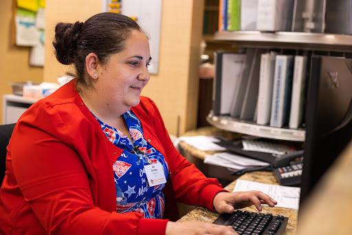 A woman in a red cardigan smiles while working at her desk, typing on a computer.