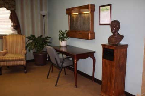 A quiet office corner featuring a wooden desk, chair, bust on a pedestal, plants, and a floor lamp.