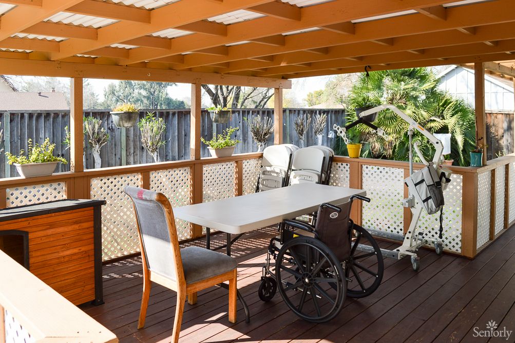 A sunny patio with a table, chairs, and a wheelchair under a wooden pergola.