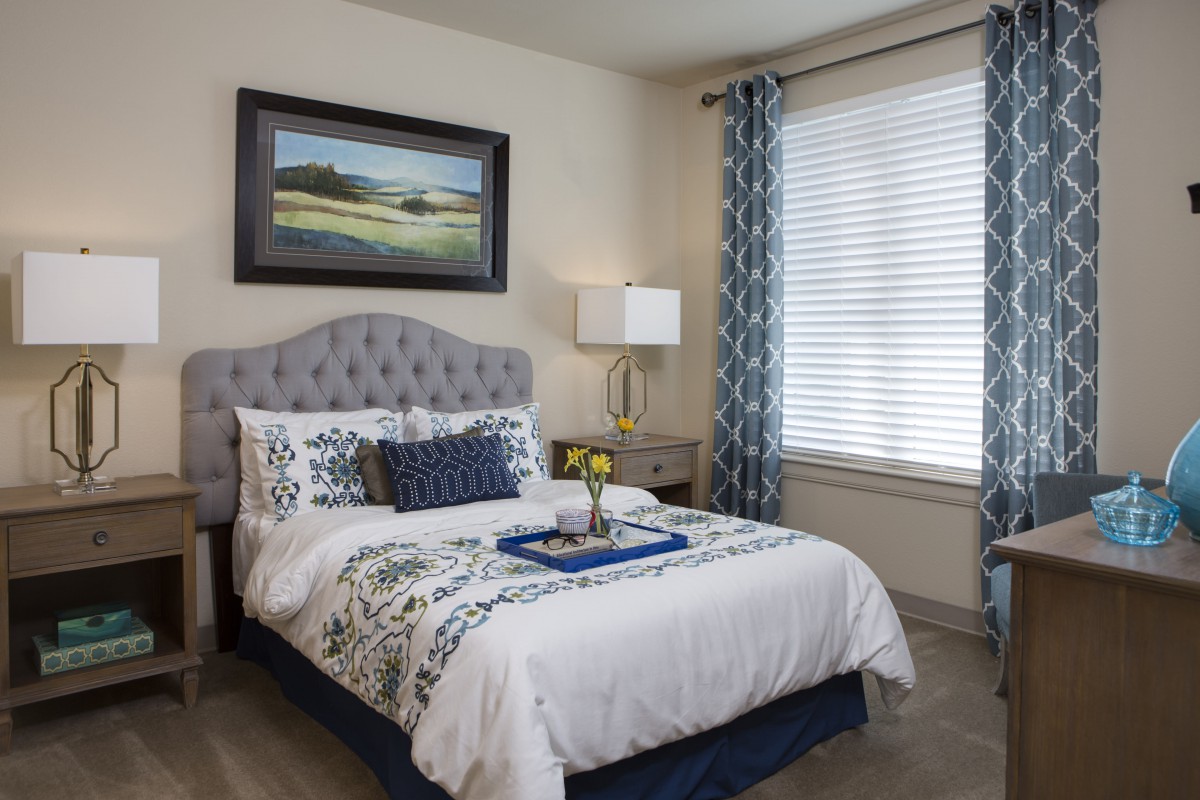 A cozy and elegantly decorated bedroom featuring a bed with patterned linens, a lamp on the nightstand, and a large window with curtains.