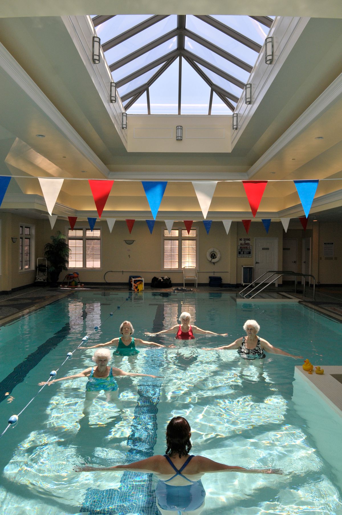 A group of seniors participate in a water exercise class in an indoor pool, illuminated by natural sunlight streaming through the skylight.