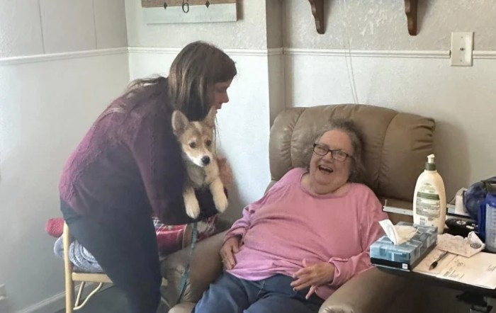 A woman holding a dog interacts with an elderly lady who is smiling in a chair.