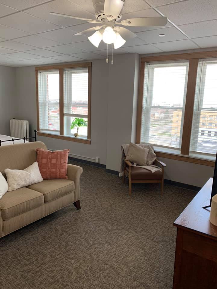 A cozy living room with large windows providing natural light, featuring a sofa, armchair, TV, and indoor plant.
