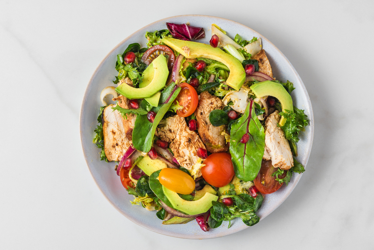 A colorful salad with grilled chicken, avocado, cherry tomatoes, mixed greens, and pomegranate seeds.