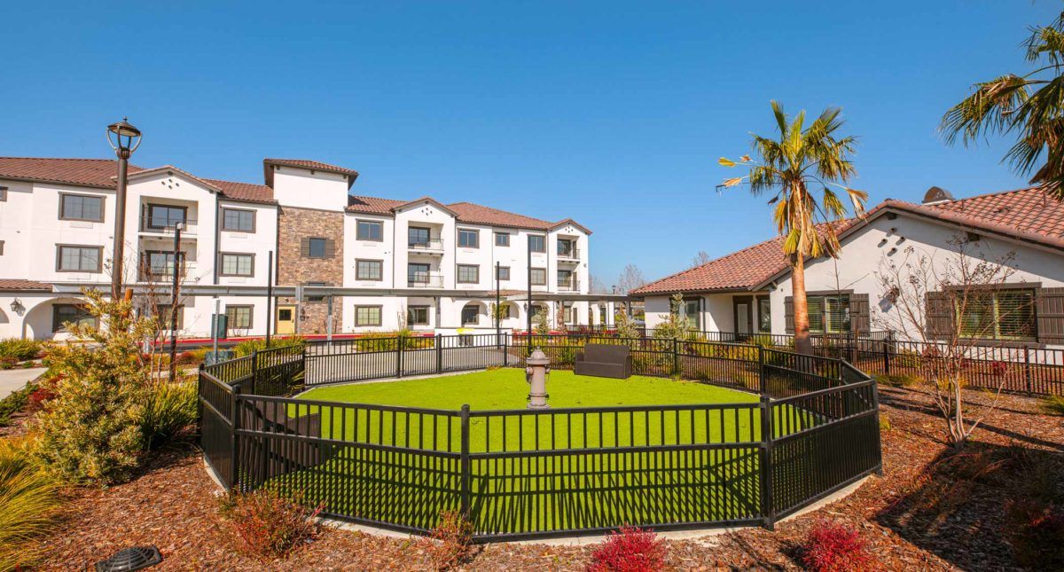 A well-maintained apartment complex with a green lawn and a fenced area under a clear blue sky.