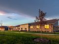 A modern building with illuminated windows, surrounded by a grassy landscape and a small tree at dusk.
