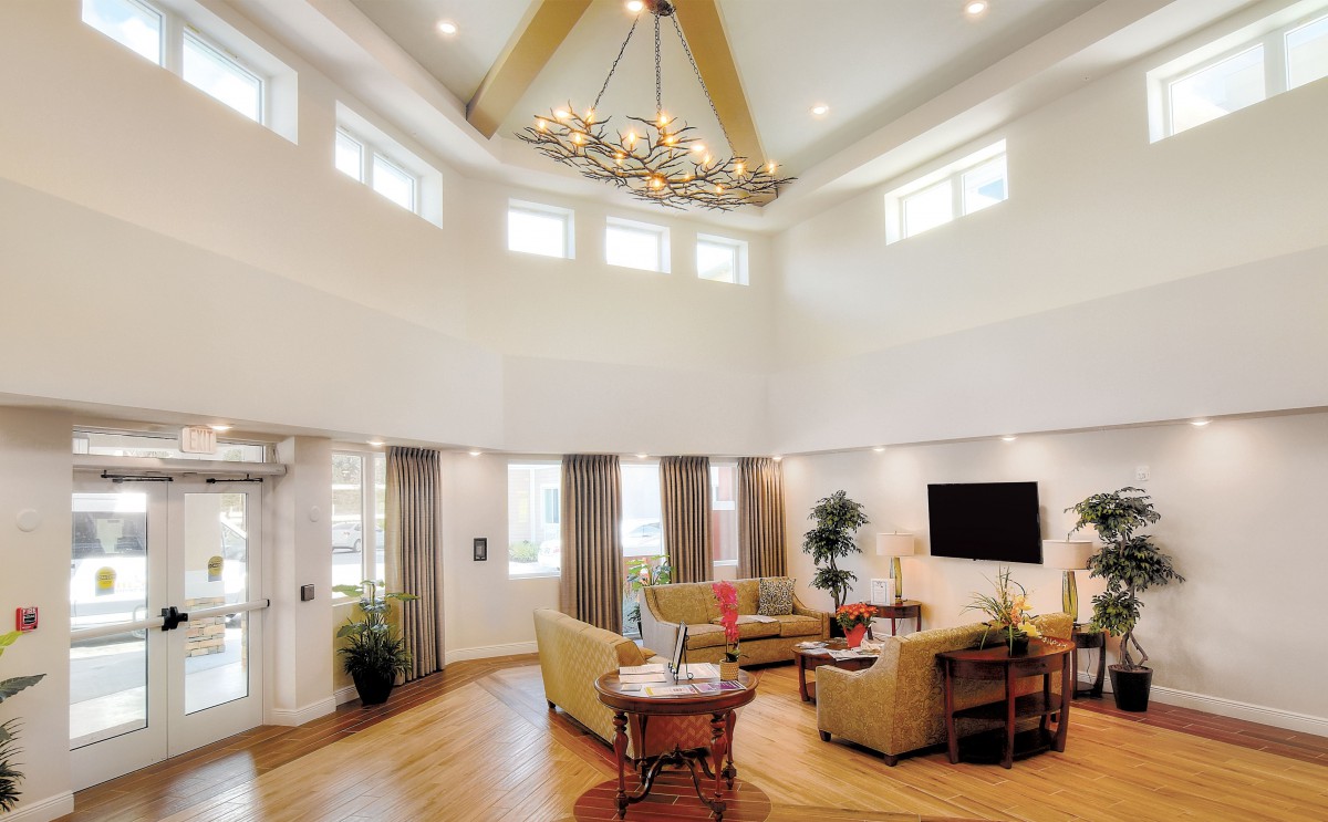 A spacious and elegantly designed living room featuring a high ceiling with a chandelier, modern furniture, and ample natural light from multiple windows.