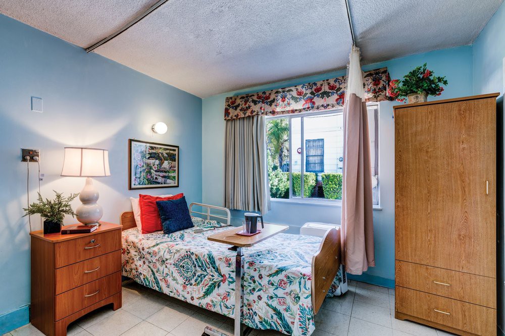 A cozy hospital room with a single bed, floral bedding, and a large window allowing natural light.