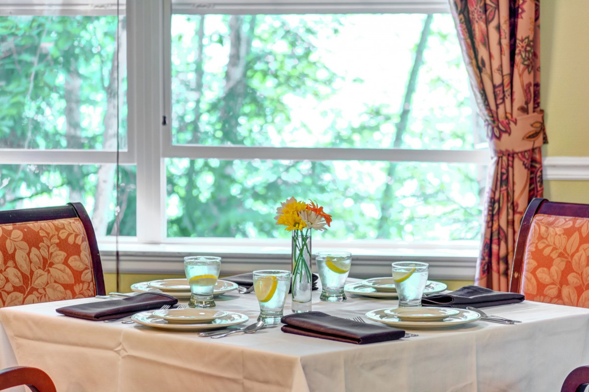 An elegantly set dining table with glasses, plates, and a flower centerpiece by large windows.