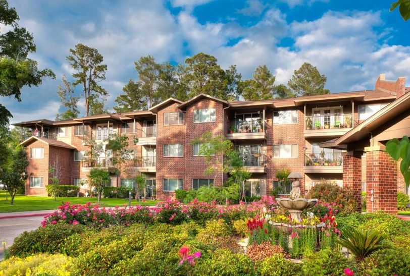 A scenic view of an apartment building surrounded by vibrant flowers and trees under a cloudy sky.