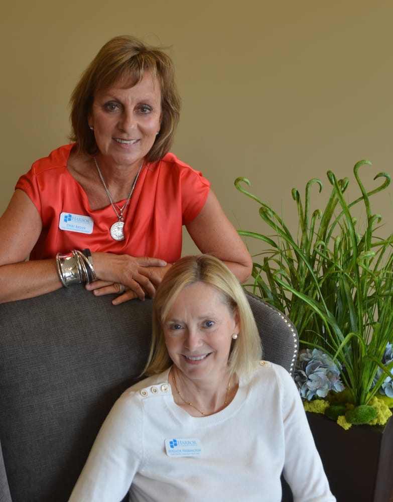 Two women smiling indoors, with one sitting and the other leaning on a chair beside some plants.