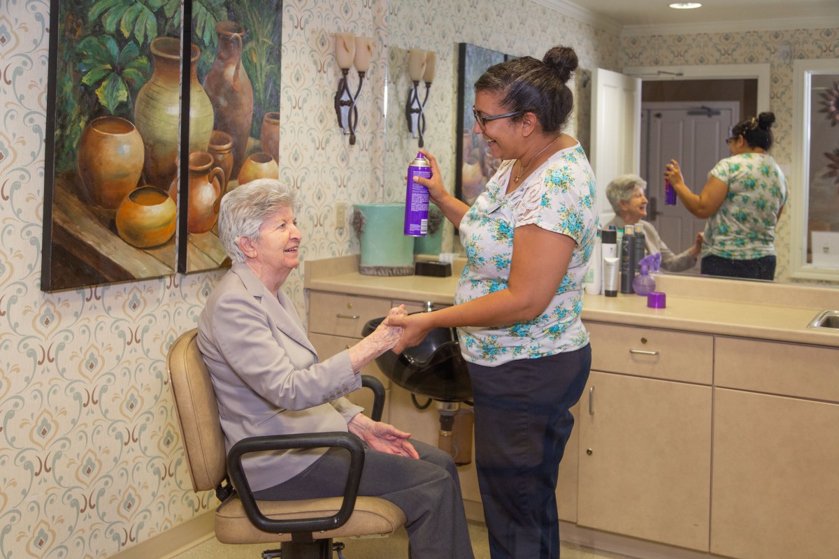 A hairstylist assists an elderly woman at a hair salon, holding a bottle of hair spray.