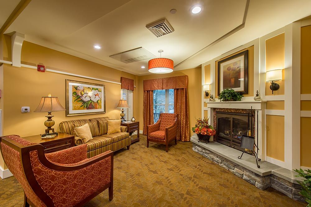 A cozy living room with warm lighting, featuring a fireplace, armchairs, and a sofa adorned with artwork on the walls.