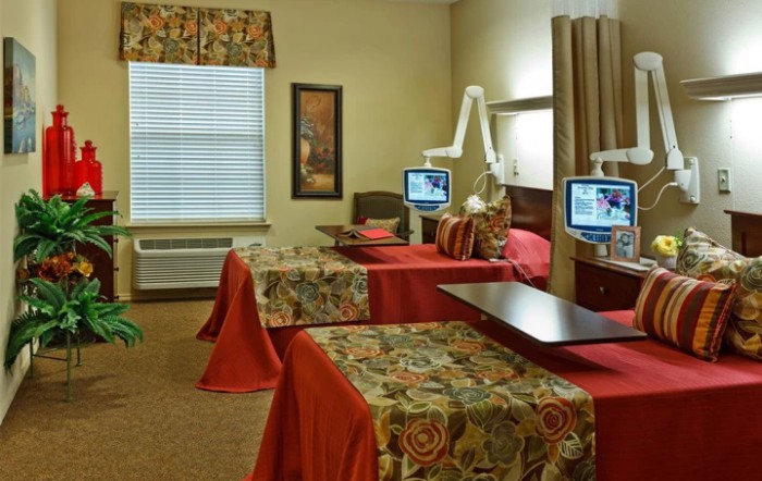 A comfortable and well-equipped nursing home room with two hospital beds, floral bed covers, and individual TVs for each bed.