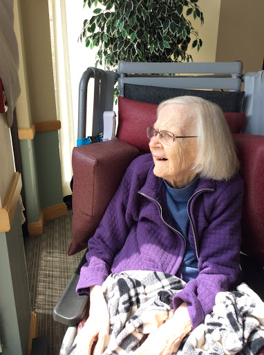 An elderly woman smiles while seated in a comfortable chair, enjoying the view from a care facility.