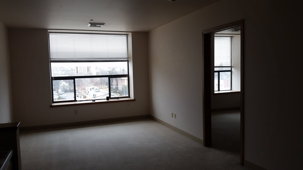 A vacant apartment room with large windows and closed blinds, featuring a carpeted floor.