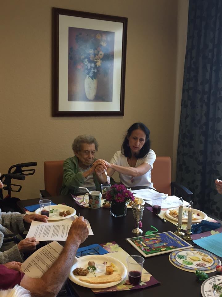 A group of people sharing a meal indoors, with one person assisting an elderly individual.