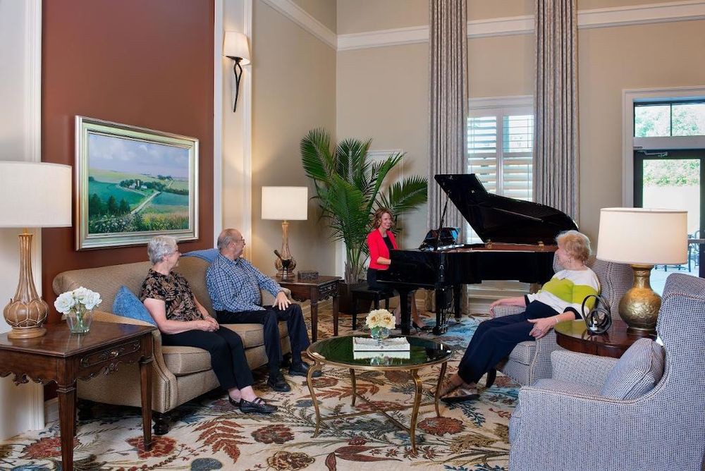 A cozy living room with people gathered around a grand piano, engaged in conversation.
