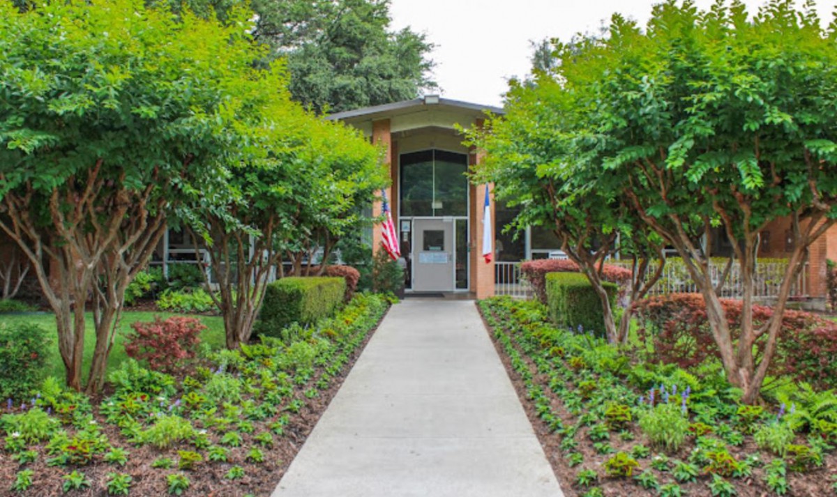 A pathway lined with trees and shrubs leading to a building entrance adorned with a flag.