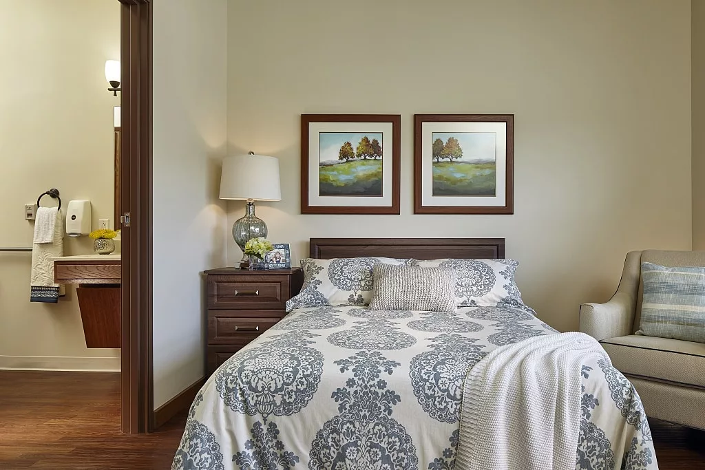 A cozy bedroom featuring a neatly made bed with decorative pillows and patterned bedding, flanked by two framed pieces of artwork on the wall and a bedside table with a lamp.