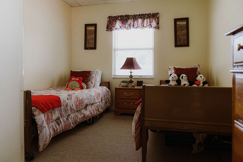 A cozy bedroom featuring twin beds adorned with stuffed animals, a central window with a patterned valance, and a bedside lamp.