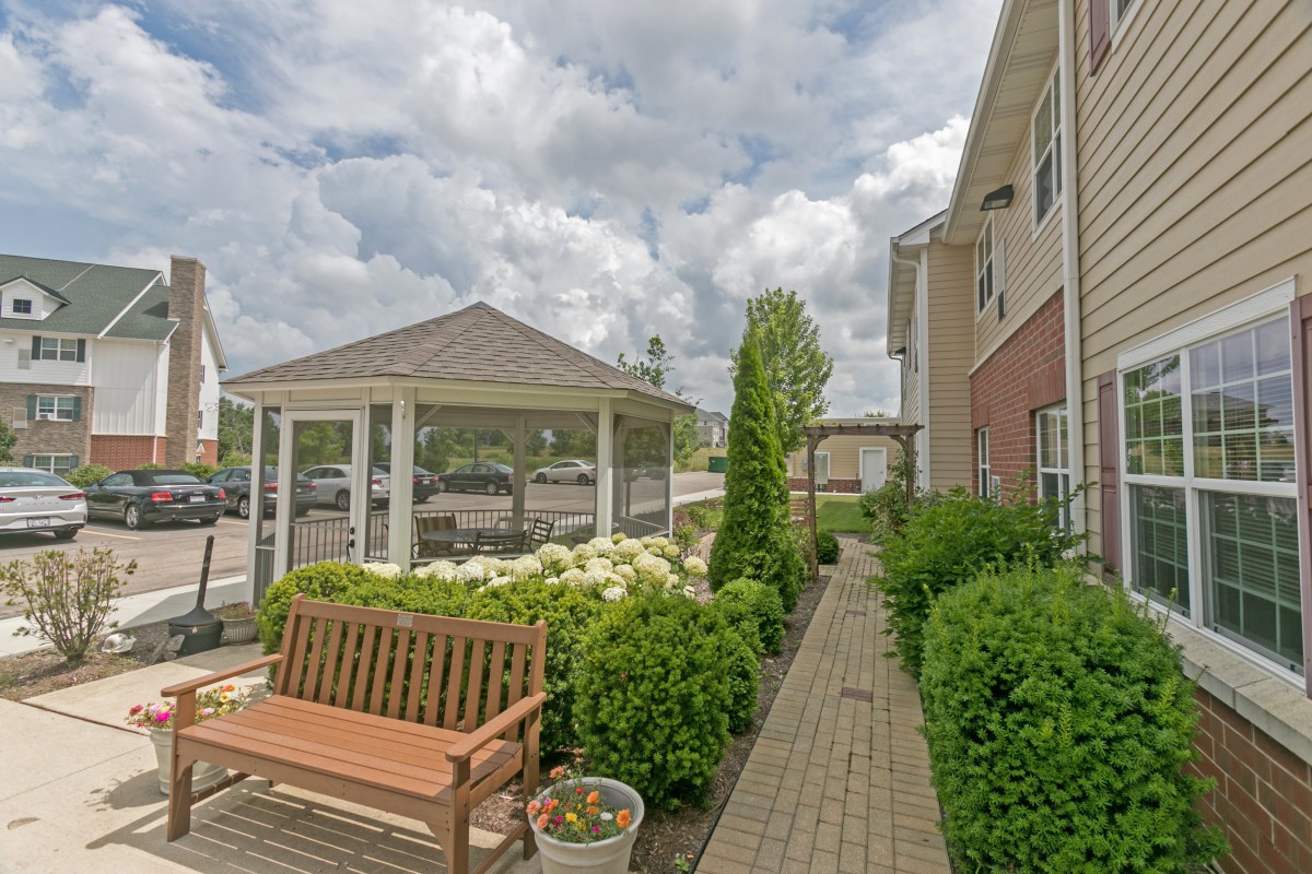 A peaceful outdoor setting featuring a gazebo, bench, and a brick path lined with well-maintained shrubs under a cloudy sky.