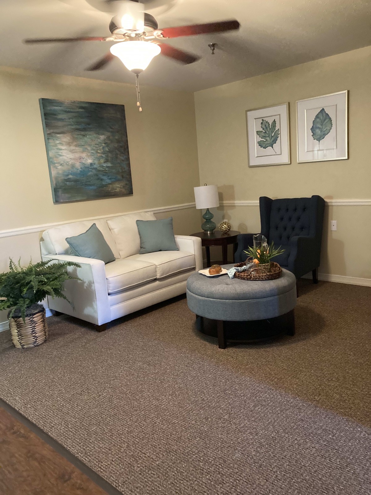 A cozy living room with a white sofa, blue armchair, and matching ottoman, adorned with artwork and an indoor plant.