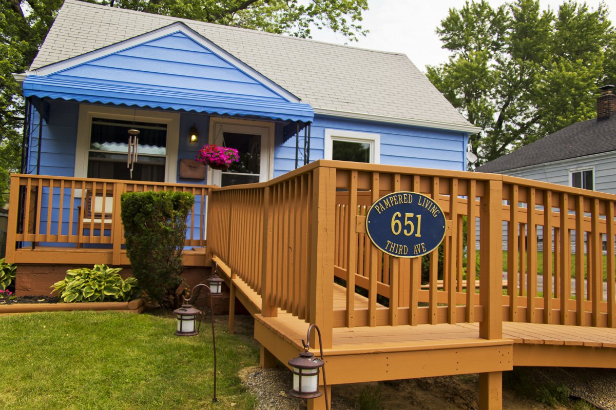 A cozy blue house with a wooden front porch and address sign, surrounded by a small garden.