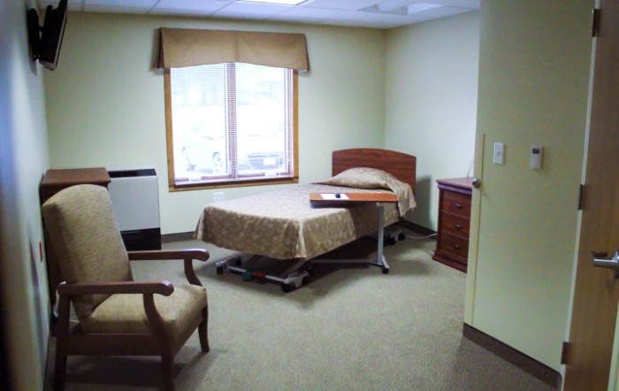 A simple and clean hospital or care facility room with a single bed, chair, and window.