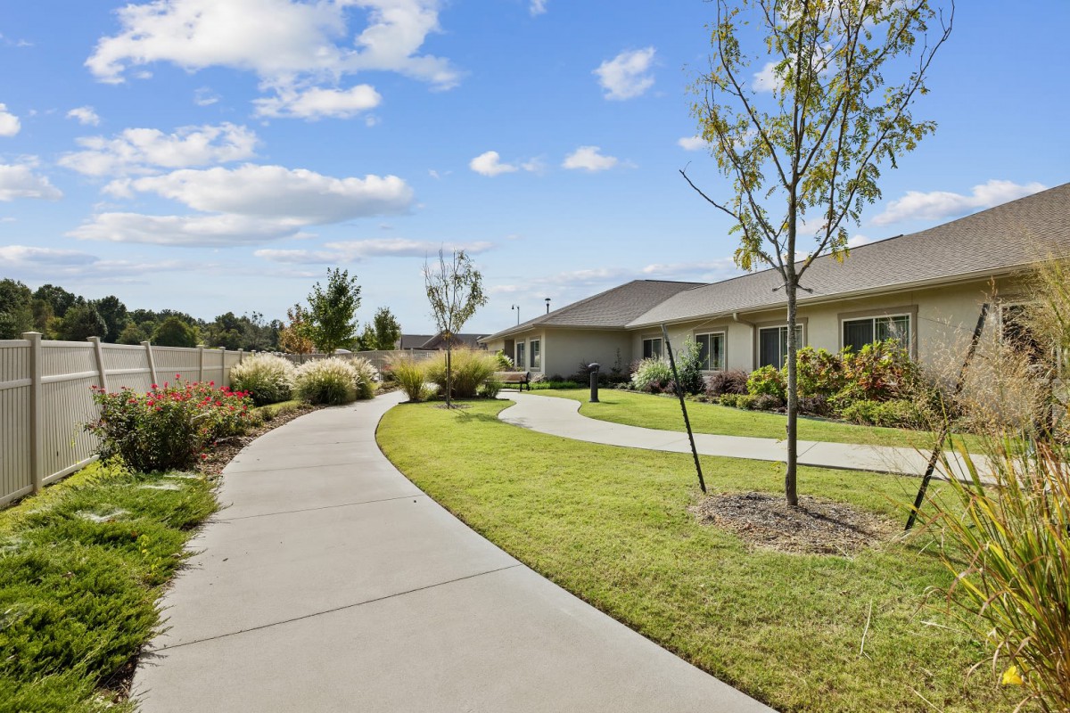 A scenic view of a building with a winding pathway, surrounded by well-maintained landscaping and trees under a blue sky with clouds.