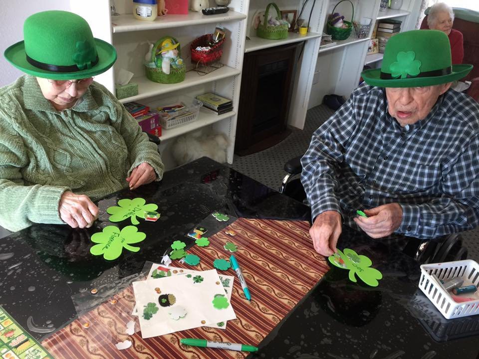 Two people wearing green hats enjoy crafting clovers for St. Patrick's Day.