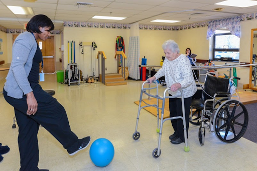 An elderly woman using a walker participates in an exercise activity with the help of a caregiver, while a blue ball is placed on the floor.