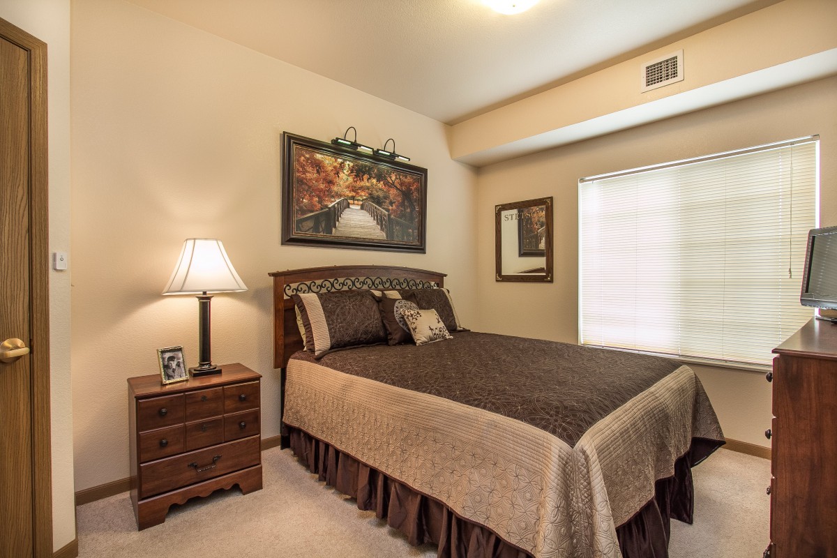 A cozy bedroom with warm lighting, featuring a neatly made bed, a wooden nightstand with a lamp, and artwork on the walls.