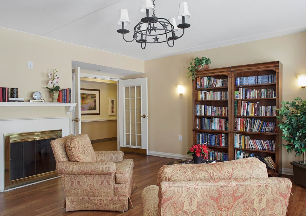 A cozy living room featuring comfortable chairs, a large bookshelf filled with books, a classic chandelier, and a fireplace, all set on a polished wood floor.