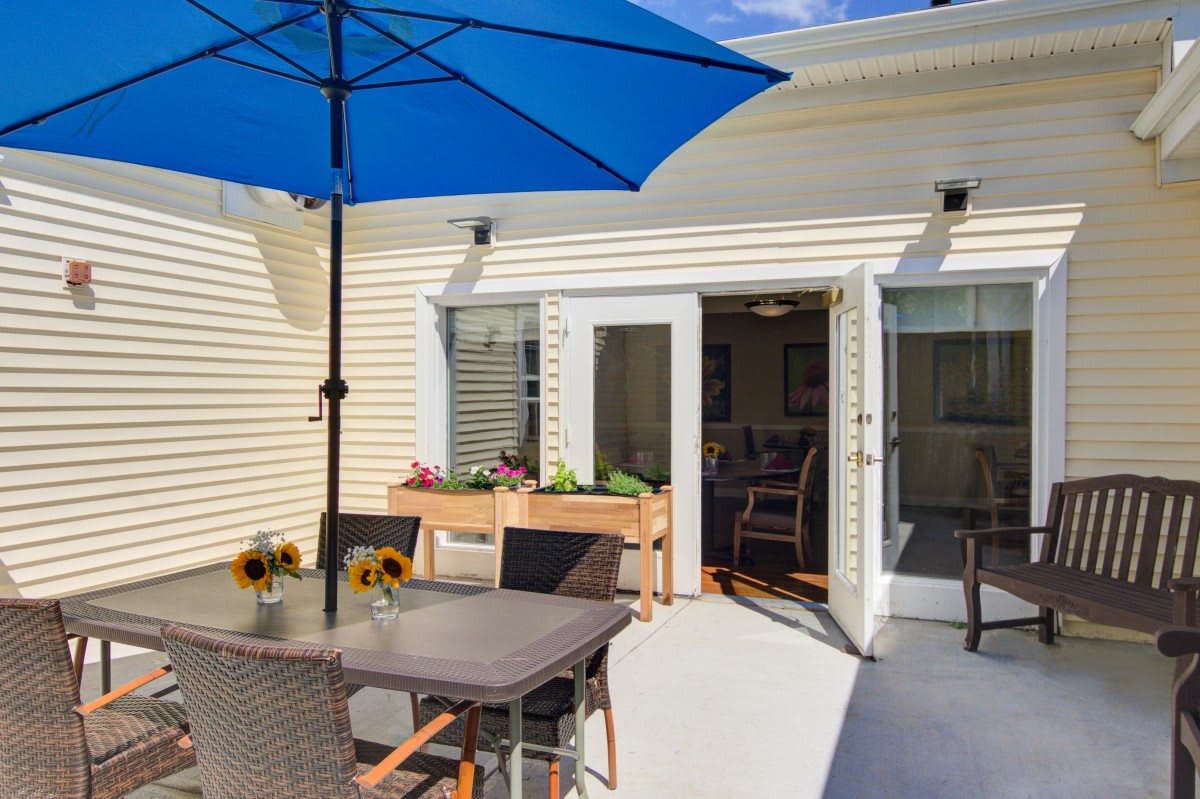 A sunny patio with a dining table, blue umbrella, and sunflowers by sliding doors.