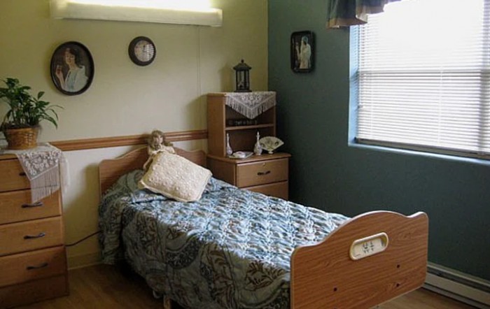 A cozy bedroom featuring a single bed with a quilted blanket, a small shelving unit with decorative items, and a window letting in natural light.