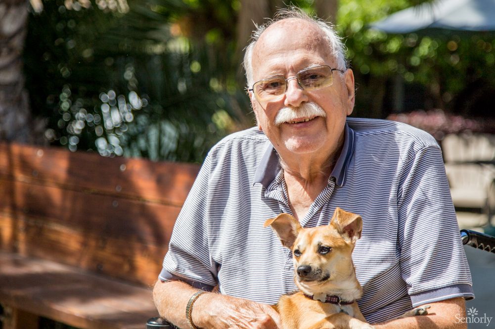 An elderly man smiling while holding his small dog outside.