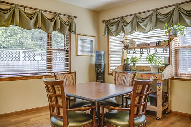 A cozy dining area with a wooden table and chairs, surrounded by windows with curtains and adorned with indoor plants.