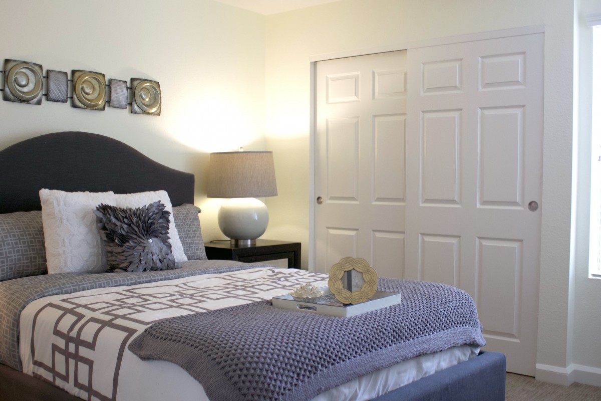 A neatly arranged bedroom with a bed, side table, and lamp, featuring decorative pillows and a closet in the background.