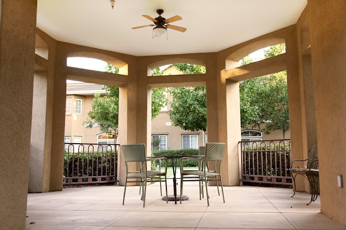 A cozy patio with a table and chairs, shaded by a ceiling fan, overlooking an apartment complex.