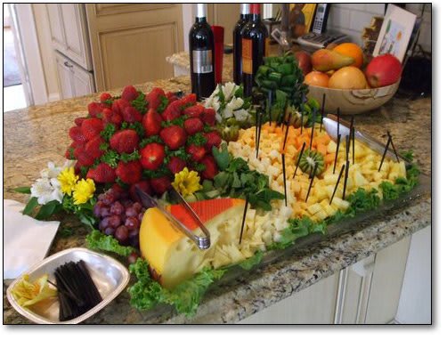 A beautifully arranged display of cheeses, strawberries, and grapes accompanied by wine bottles on a buffet table.