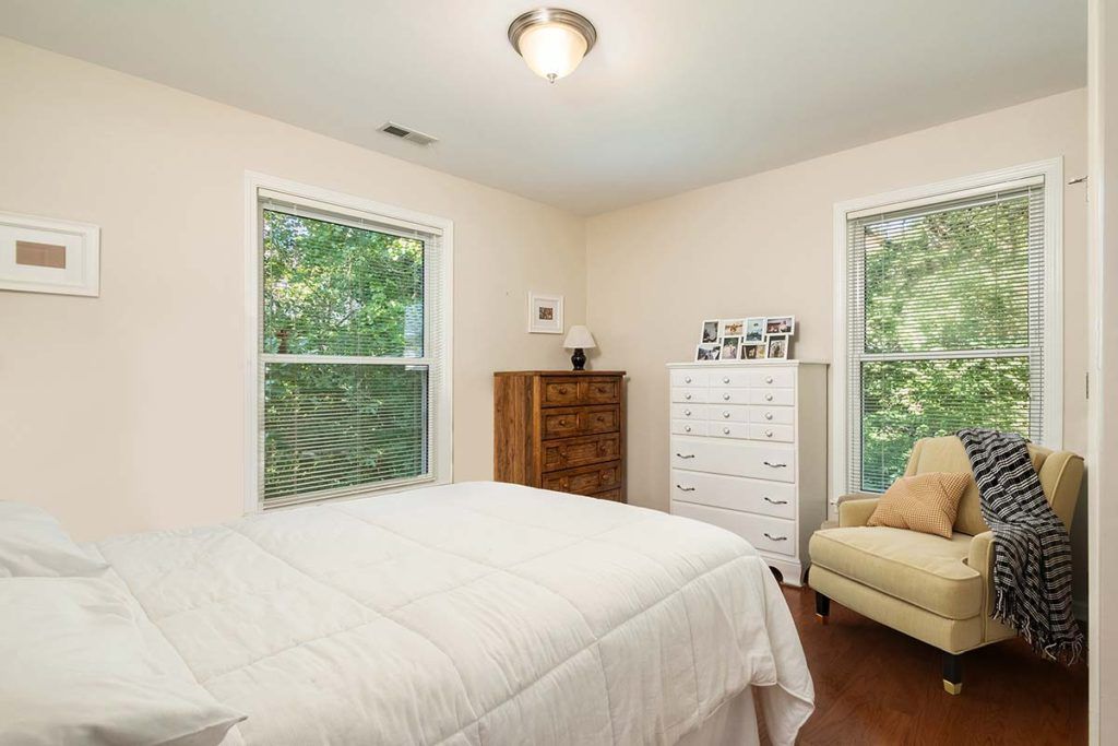 A cozy bedroom featuring two large windows, a bed with white bedding, two chests of drawers, and a yellow chair on a hardwood floor.