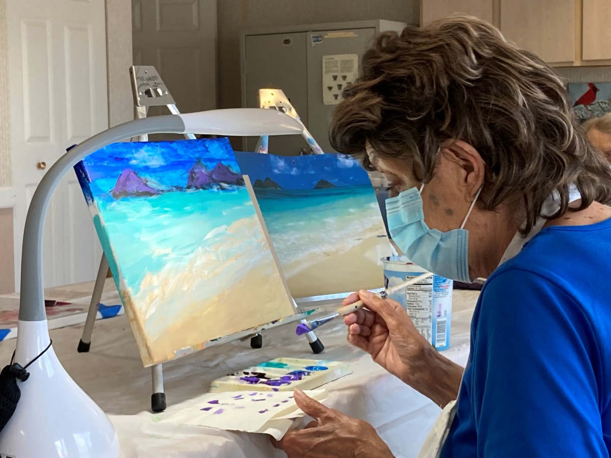 An artist wearing a mask carefully paints a beach scene on a canvas indoors.
