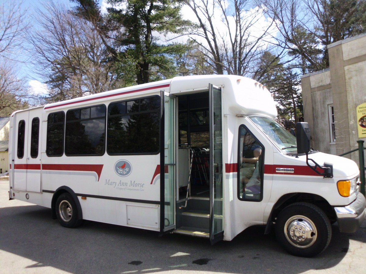 A white shuttle bus with an open door parked by the roadside and surrounded by trees.
