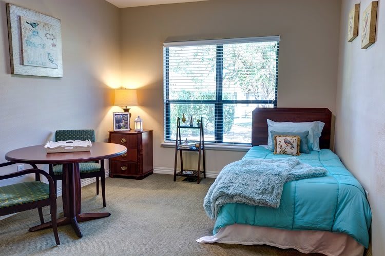 A cozy bedroom with a neatly made bed, a small table and chair, and a lamp by the window.