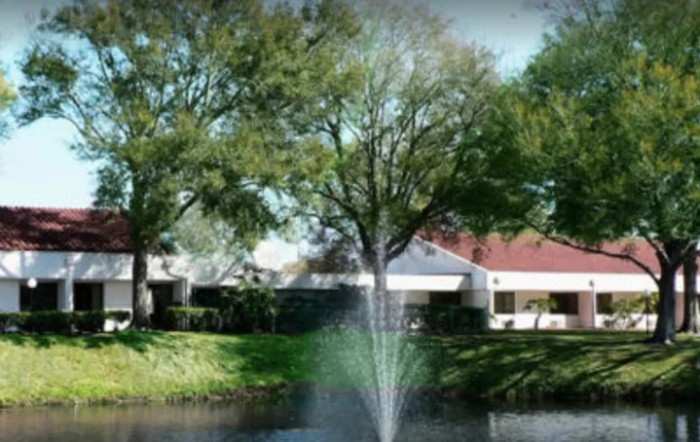 A serene view of a fountain in a pond with surrounding trees and buildings.
